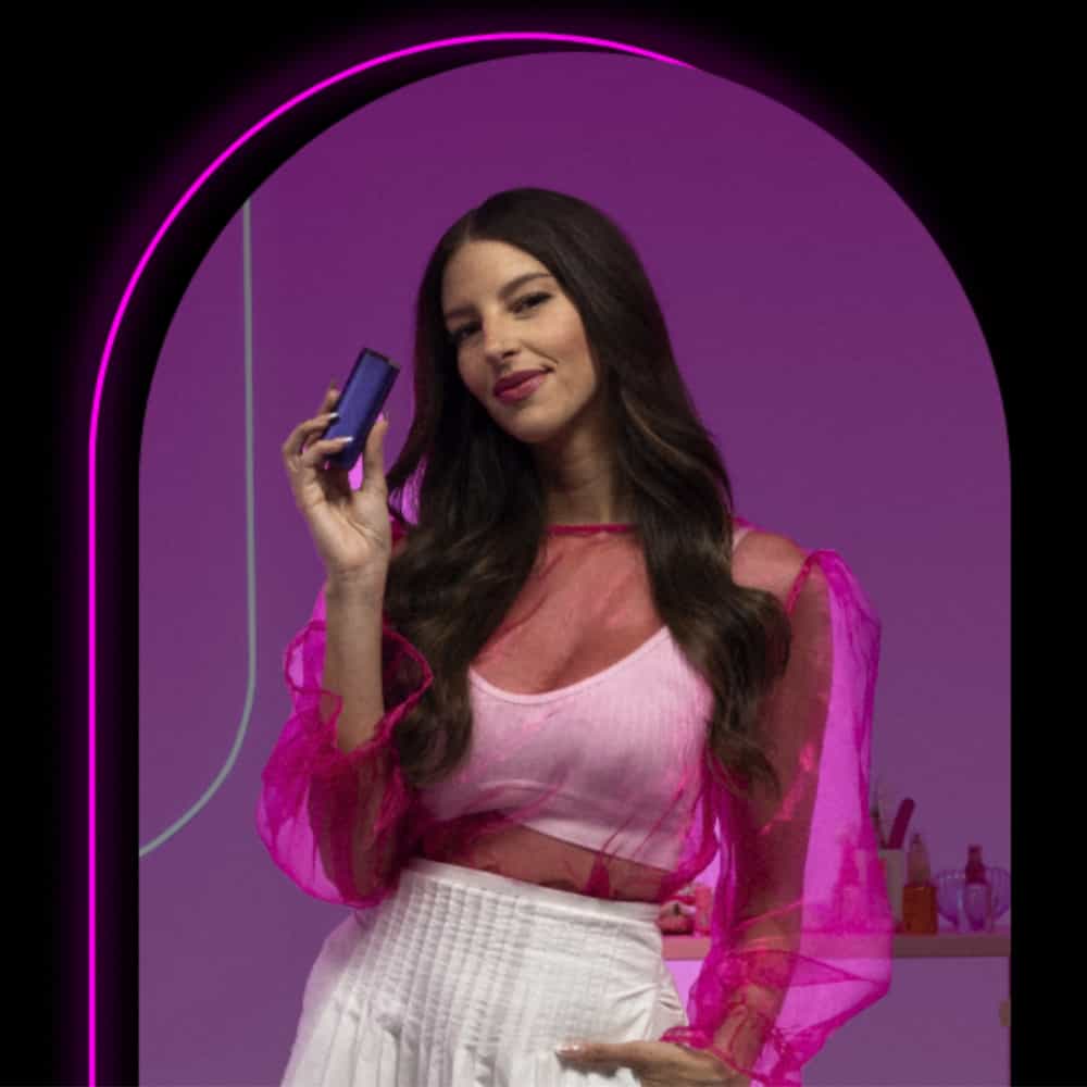 A woman with long brown hair wears a sheer pink top and white skirt, smiling and holding a small rectangular device. The background is purple with a glowing neon border around the image.