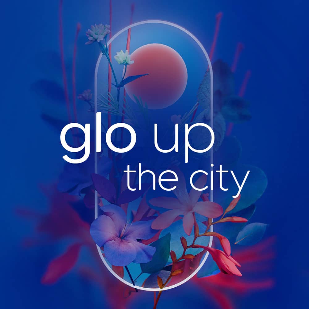 A neon oval frame with tropical flowers and foliage inside, set against a blue and pink background. White text in the center reads, glo up the city.